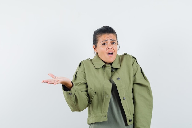 Woman spreading palm aside in jacket, t-shirt and looking confused