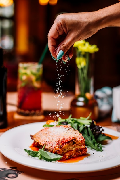 Woman sprinkles grated parmesan particles on lasagne