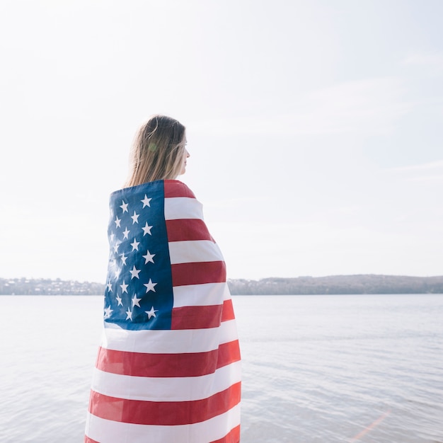 Woman standing completely wrapped in American flag and looking at sea