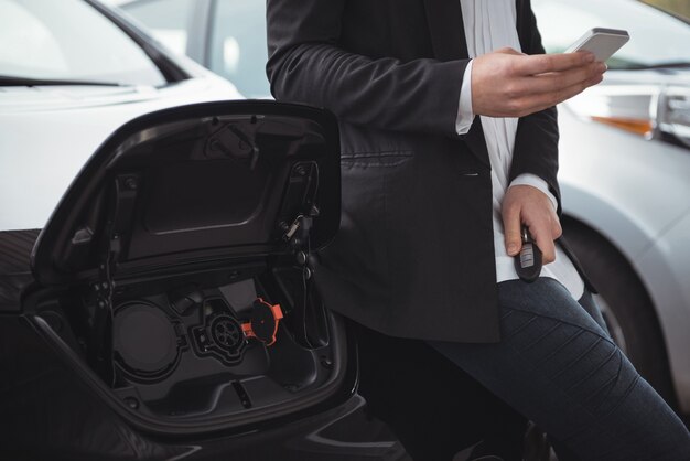 Woman standing next to electric car and using mobile phone