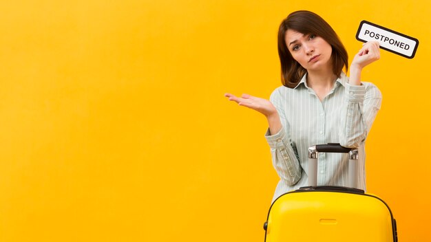 Woman standing next to her luggage while holding a postponed sign with copy space