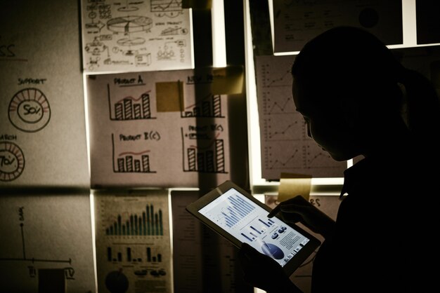 Женщина, стоящая рядом с окном, покрытым бизнес-диаграммами и работающим на планшете
