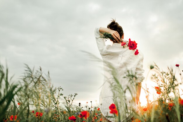 Woman stands holding a poppies flower bouquet over a back, among the meadow