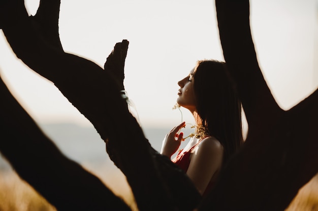 Woman stands behind an old tree on the field watching the sunset