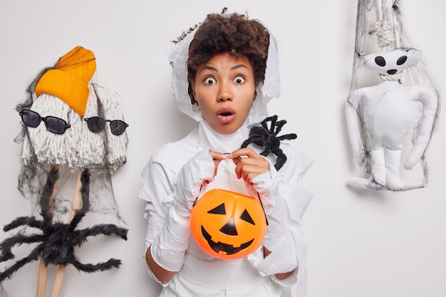 woman stares bugged eyes holds carved pumpkin and spooky spider poses on white with creepy creatures around. Halloween decor