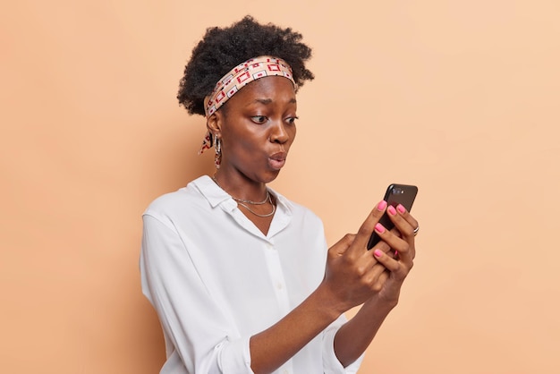 woman stares at smartphone screen has shocked expression reads news dressed in white shirt and headband isolated on beige makes online shopping. Technology concept