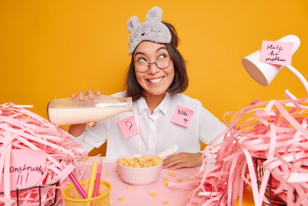 woman starts her day with delicious healthy breakfast eats cereals with milk looks away with happy dreamy expression wears sleepmask white shirt and spectacles makes notes with list to do