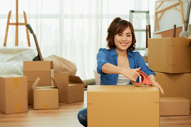 Woman sticking adhesive to the package box smiling at camera seated on the floor