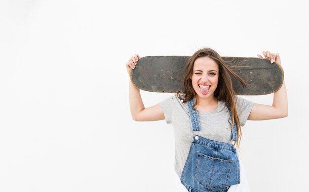 Woman sticking her tongue out holding skateboard on her shoulder against white backdrop