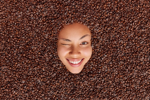 Free photo woman sticks head through brown roasted coffee beans winks eye smiles broadly expresses positive emotions