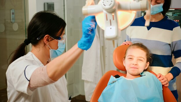 口腔病学の椅子に座っている小さな患者を検査するためにランプを点灯している女性の口腔病学者の技術者。看護師が検査のためのツールを準備している間、医者は歯の健康をチェックしている女の子に話します