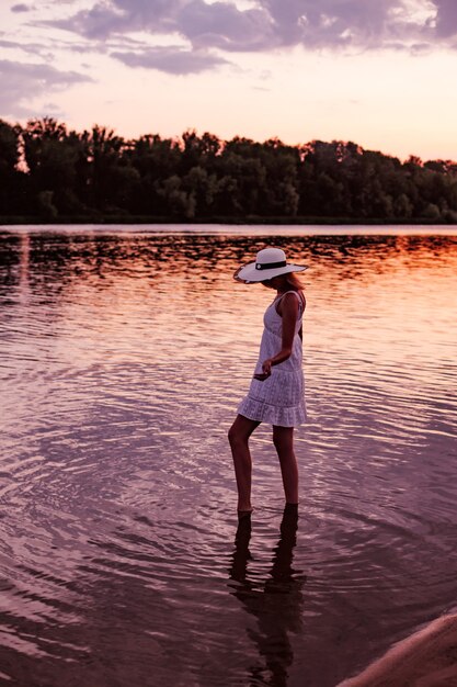 A woman in a straw hat at sunset. a beautiful young slender blonde stands in the water in a white summer dress and raises her arms to the sides.