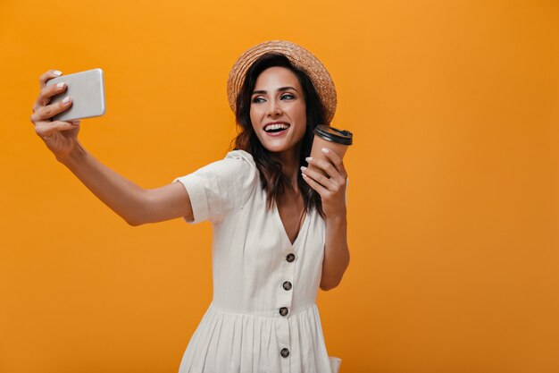 Woman in straw hat and white dress makes selfie on smartphone and holds glass of tea. Adult lady in light outfit takes photo with coffee in her hands.