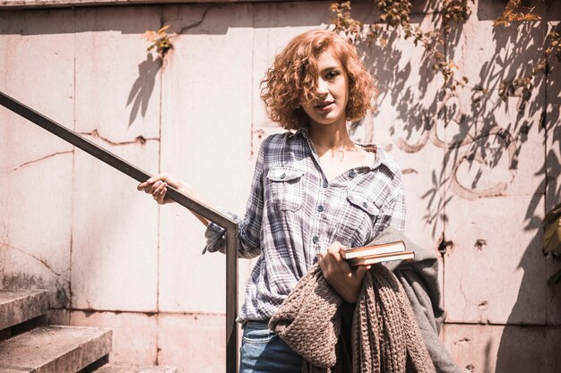 Woman in street wear with books and scarf in hand holding rail 