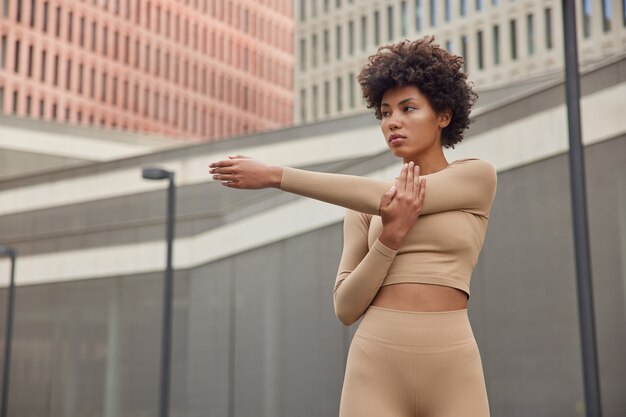 woman stretches arms dressed in tracksuit does exercises warms up before workout looks seriously into distance trains outdoor demonstrates determination