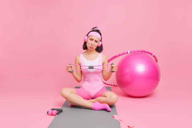 Woman stretches hands with resistance band dressed in activewear crosses legs on mat listens music via headphones surrounded by sport equipment. healthy lifestyle concept