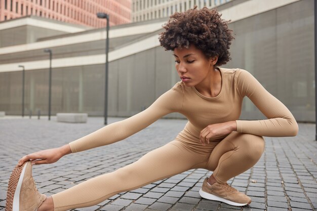 女性はハムストリングのポーズを伸ばします屋外はベージュのスポーツ服を着て、スニーカーは体調が良いトレーニングをします