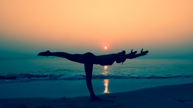ヨガで彼女の体を延伸する女性がビーチで日没でポーズ