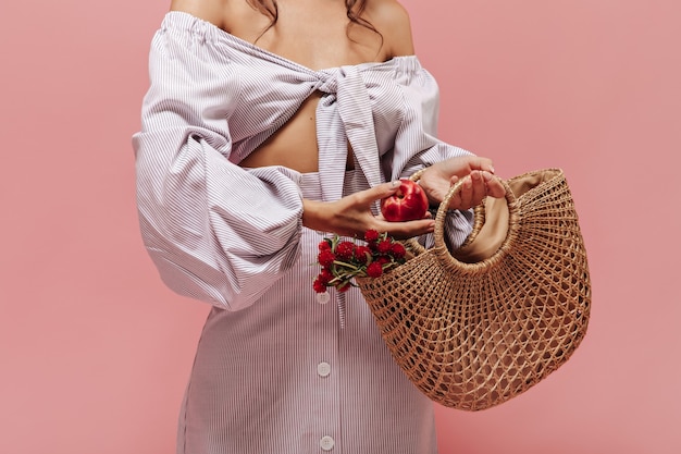 Woman in striped modern blouse and skirt with white button laying red apple in cute straw handbag with beautiful flowers