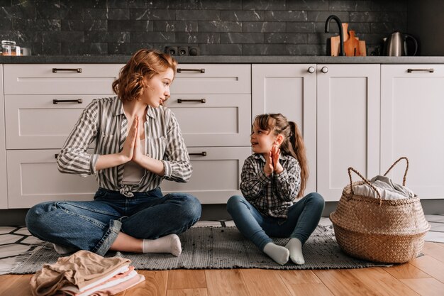 Woman in striped shirt is sitting on floor and shows her daughter how to meditate.