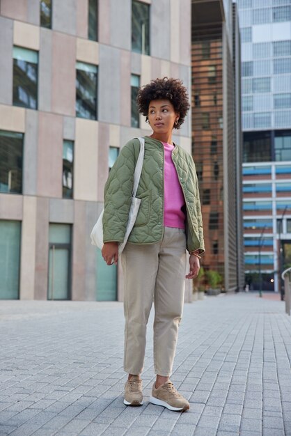 woman strolls in city against city buildings poses in centre looks away thinks about something has excursion at downtown