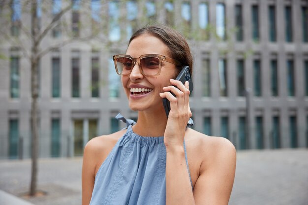 휴대 전화를 통해 현대 도시 회담의 거리를 산책하는 여성은 트렌디 한 선글라스를 착용하고 파란 드레스 미소는 긍정적 인 대화 중에 로밍 연결 웃음을 광범위하게 사용합니다