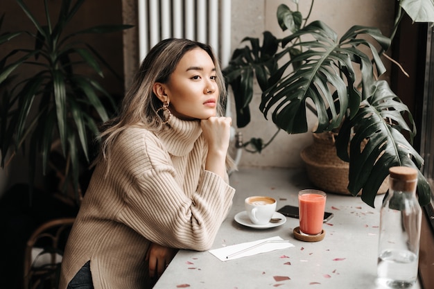 woman in stylish beige sweater looks thoughtfully into distance, leaning on table with cup of coffee and fresh juice