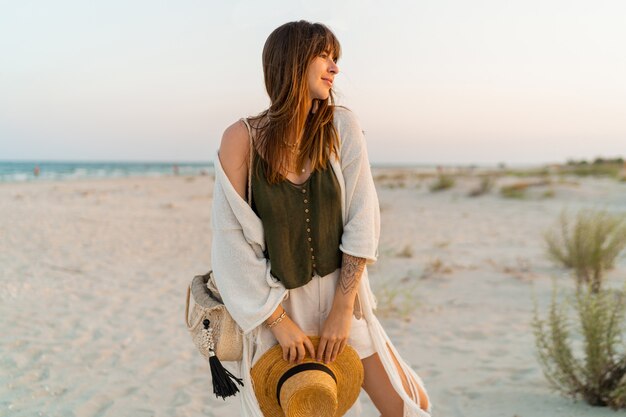 woman in stylish boho outfit holding straw bag and hat posing on tropical beach.