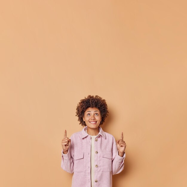 Woman in stylish clothes indicates with fore fingers above shows advertisement on top advertises promo offer poses against beige. this copy space is awesome