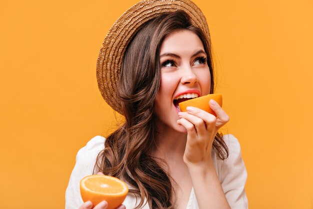 Woman in stylish hat bites delicious orange and poses on orange background.