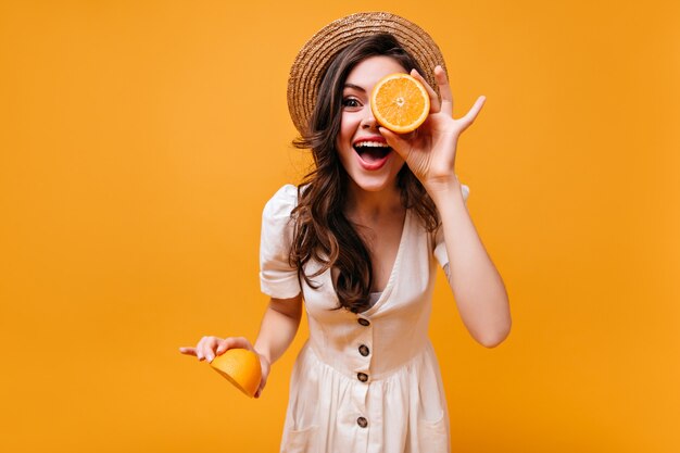Woman in stylish white dress and straw hat laughs and holds slices of oranges.