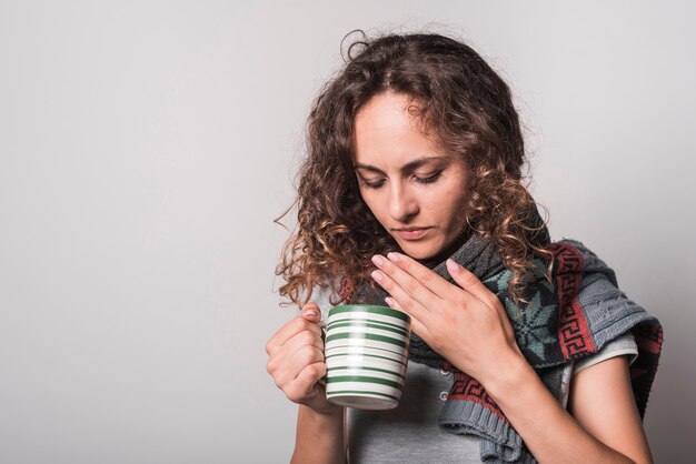 Woman suffering from cold holding coffee mug against gray background