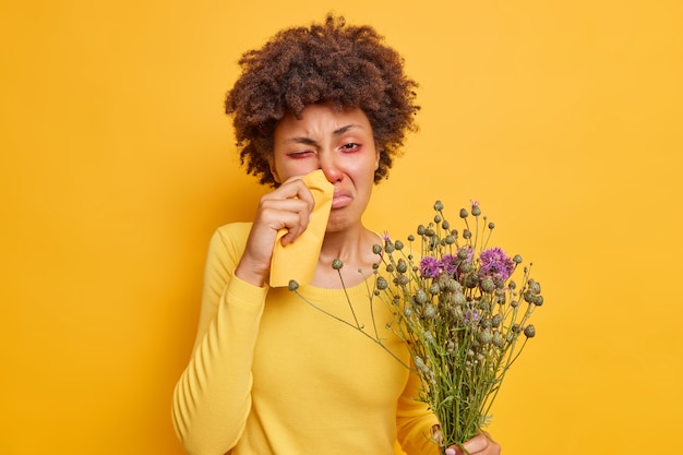 アレルギー性鼻炎に苦しむ女性がナプキンで鼻をこすり、野花のブーケを握り、鮮やかな黄色で体調不良のポーズをとる