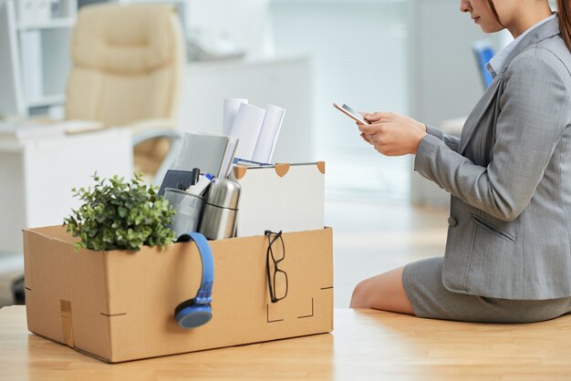 Woman in suit sitting on desk in office with belongings in box and using smartphone