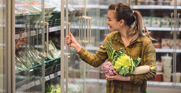 Donna al supermercato. bella giovane donna tiene in mano verdure biologiche fresche e apre il frigorifero al supermercato. il concetto di mangiare sano. raccolto