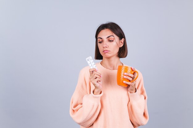 Woman in sweater on gray wall holds mug and pills with unhappy sad expression on face