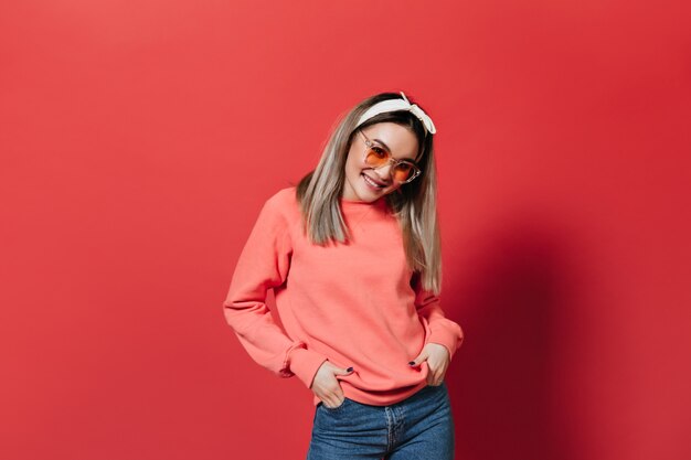 Woman in sweatshirt and orange glasses smiles sweetly on red wall