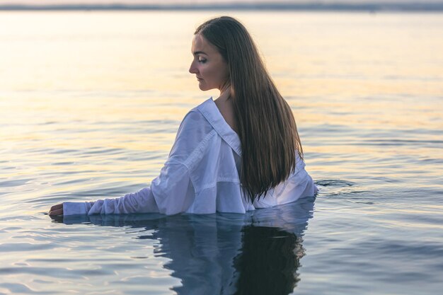 Женщина в купальнике и белой рубашке в море на закате