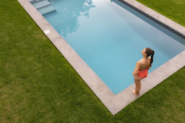 裏庭のプールサイド近くの腰に手で立っている水着の女性