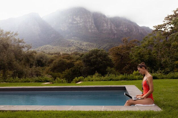 Женщина в купальниках, используя ноутбук возле бассейна на заднем дворе