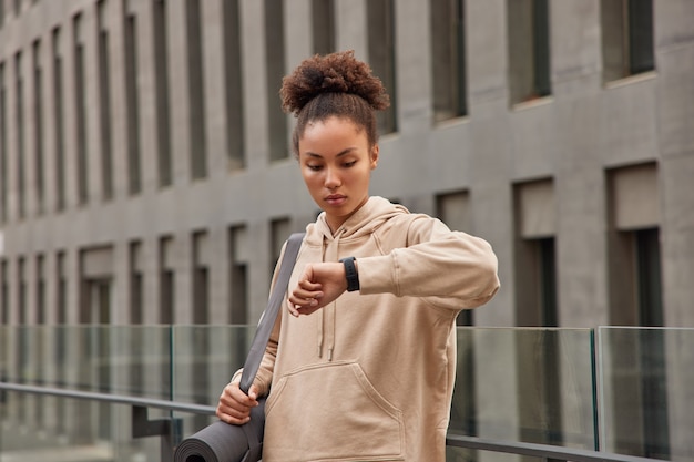 женщина делает перерыв после того, как фитнес-активность проверяет результаты на умных часах, несет свернутый карем, одетый в толстовку с капюшоном, стоит на фоне современного здания