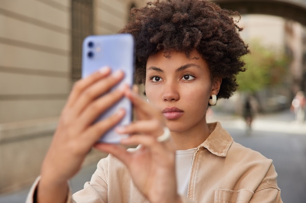 女性はソーシャルネットワークで共有するためにスマートフォンで自分の写真を撮りますカメラを注意深く見て街でレクリエーション時間を楽しんでいます