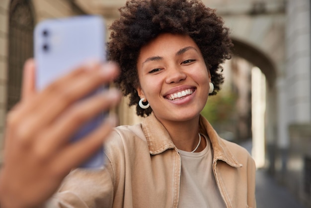 女性はスマットフォンを介して自分撮りをします携帯電話のウェブカメラで写真をクリックしますソーシャルネットワークで公開するための写真を作成します日中の路上でカジュアルなアパレルポーズを着ます