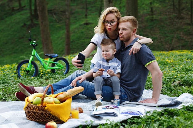 Woman takes a selfie with her parents and son sitting on a plaid in the park