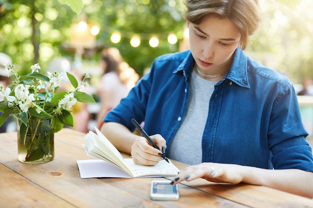 スマートフォンを使用してメモを取る女性。彼女のノートに書いている若い女性の屋外の肖像画
