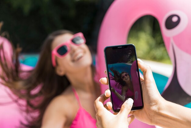 Woman taking photo laughing young female on inflatable flamingo in pool