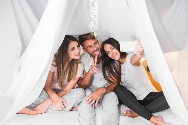 Woman taking selfie with her friends sitting on bed under the white curtain