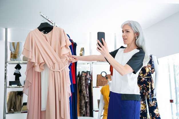 Woman talking on cell in fashion store and showing dress on rack at frontal. Medium shot. Boutique customer or communication concept