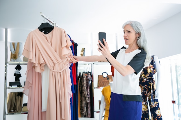 Женщина разговаривает на ячейке в магазине модной одежды и показывает платье на стойке на лобной. Средний план. Бутик-клиент или концепция коммуникации