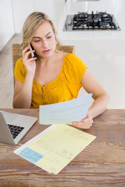 Woman talking on mobile phone while looking at bill in kitchen